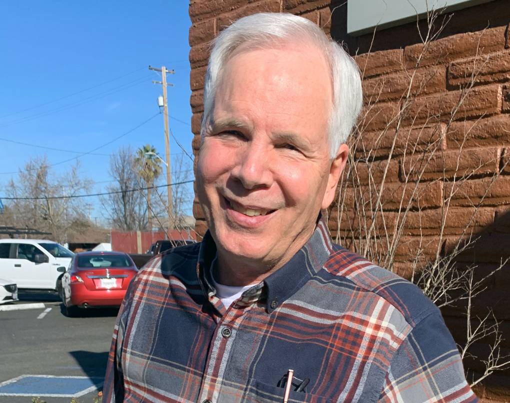 man with white hair and plaid shirt stands outside in front of brick wall with parking lot in background