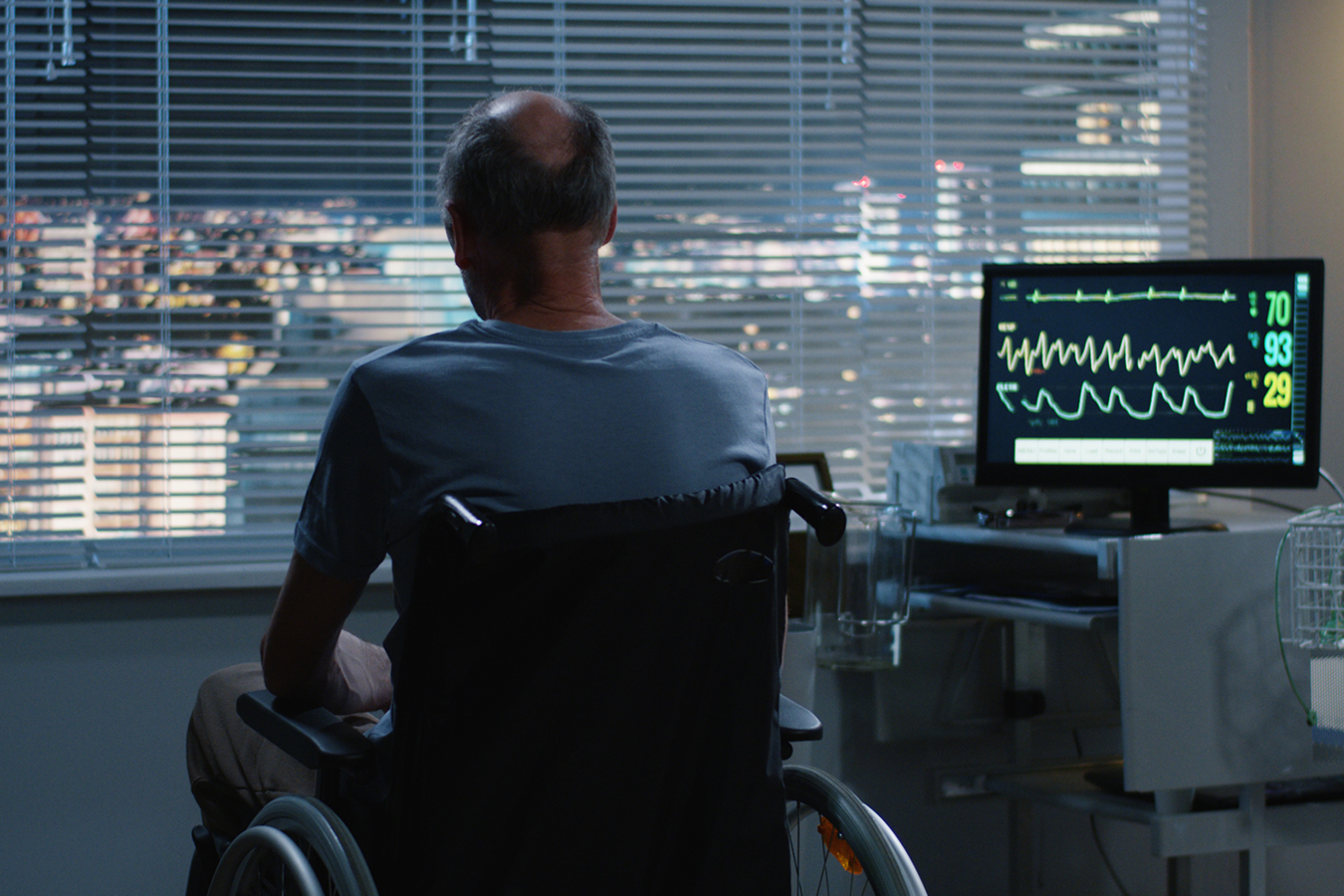 Medium shot of a elderly male patient in a wheelchair looking out a window at night