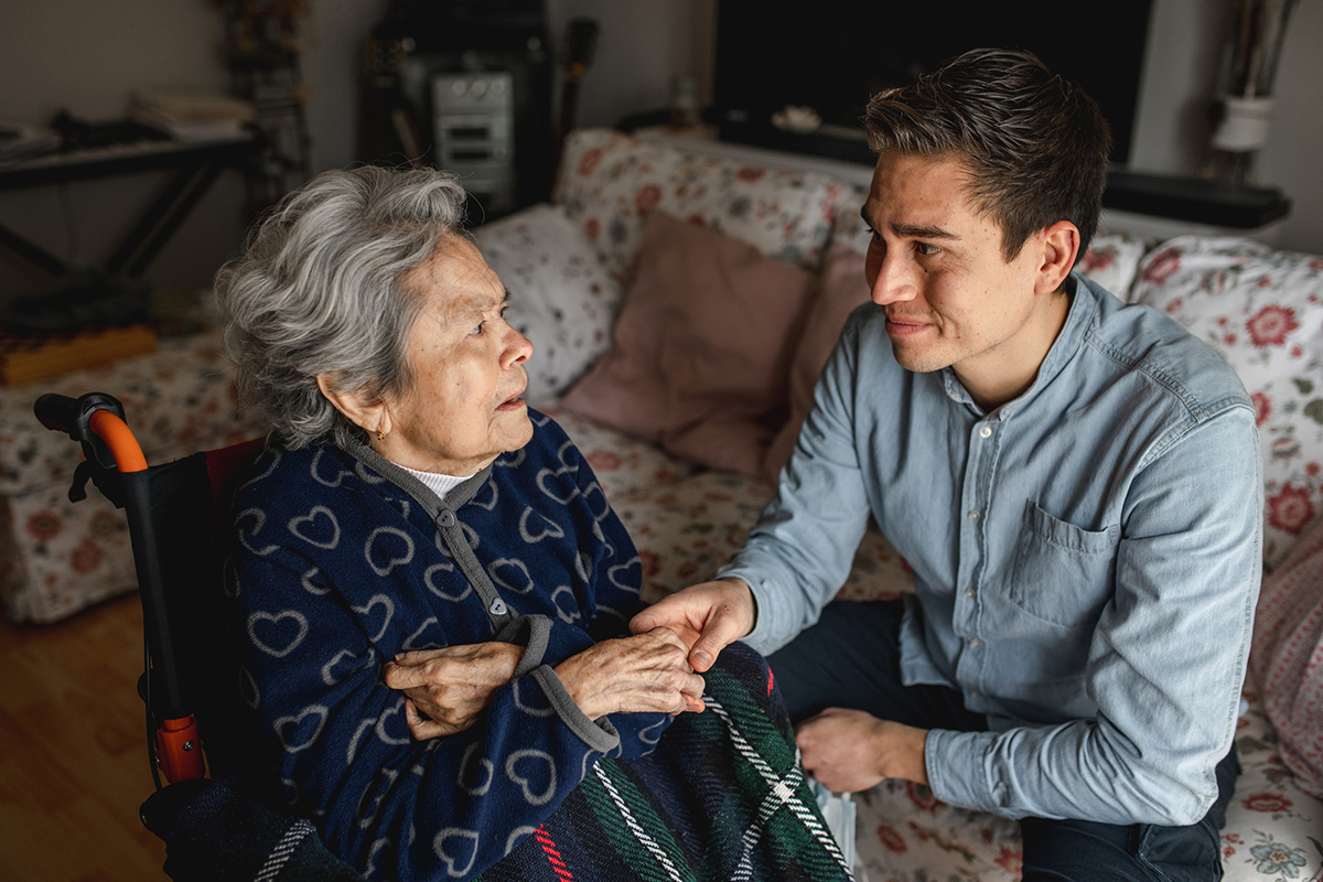 Young man sitting next to an aged woman in wheelchair, taking her hands while talking and smiling