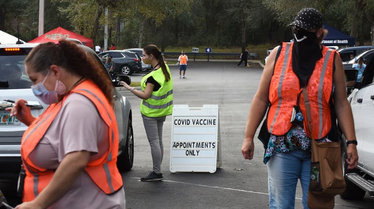 Masked workers in safety vests walk between cars in a parking lot by a sign reading COVID vaccine, appointments only.