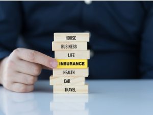 A stack of seven wooden blocks. From top to bottom, they read "home, business, life, insurance, health, car, travel." The insurance block is yellow, a man in a suit is reaching his hand towards it.