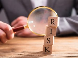 Close-up Of Wooden Blocks With Risk Word In Front Of Businessperson's Hand Holding Magnifying Glass