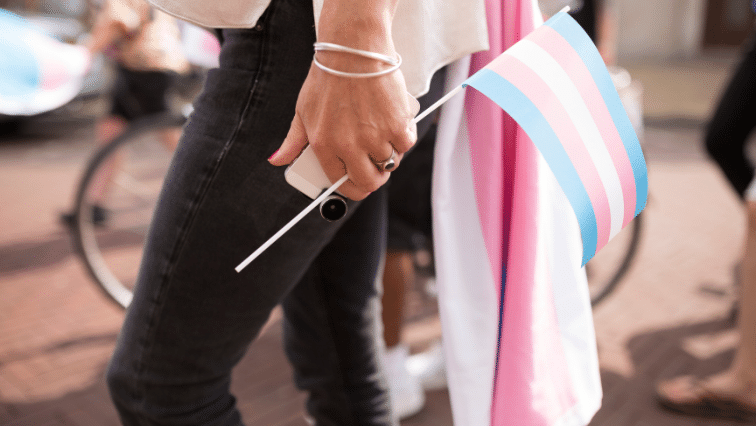 A close-up image of a person walking, holding a cell phone and a trans flag in their left hand, and a full-size trans flag draped over their shoulder.