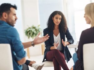 A woman in business wear sits across from another woman and a man. They are all engaged in conversation.