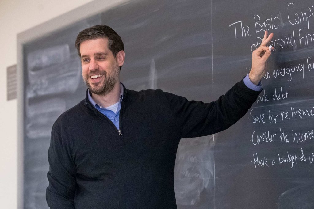 UVA associate professor Andrew Pennock, smiling writes on a blackboard: The basic components of personal finance.