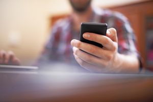 A closeup photo of a man's hand holding a smartphone. He's sitting at a table, typing on a keyboard with his other hand.