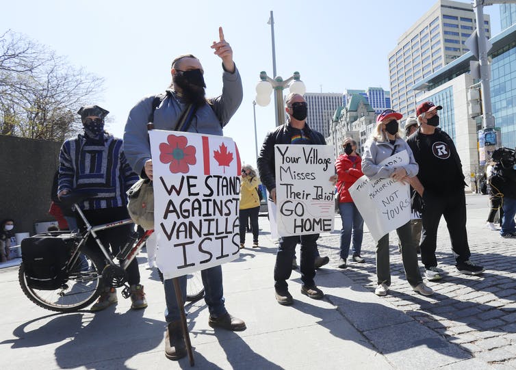 Protesters in masks carry signs, one reading 'your village misses its idiot, go home'