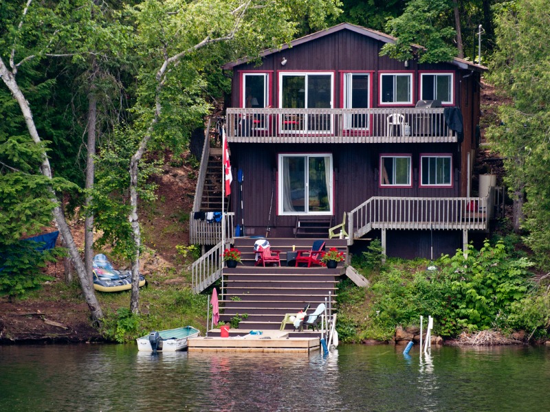 Multi-level brown cottage with white trim sits on the water and is surrounded by trees.