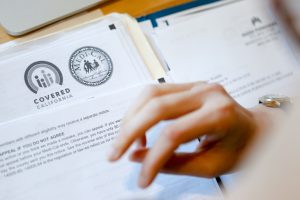 A closeup photo shows a hand flipping through a folder. At the top of a page on the left, the logos for Covered California and Medi-Cal are seen.