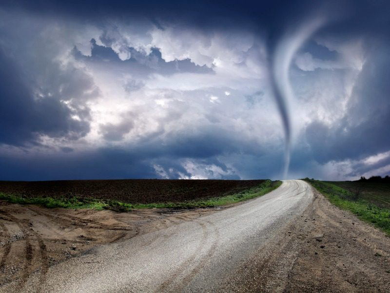 A twister is pictured at the end of a country road. The sky is dark and storming.