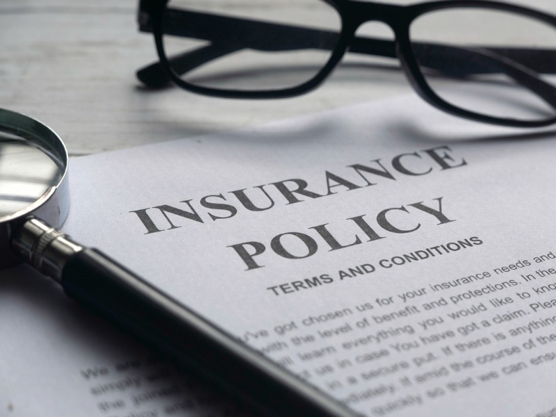 An insurance policy is pictured laying on a desk with black framed glasses and a magnifying glass atop it.