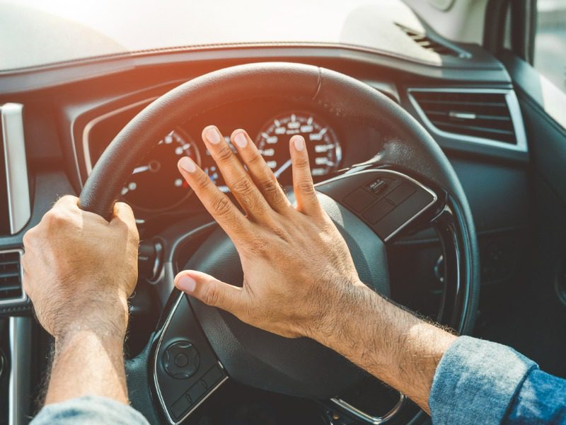 A person holds the steering wheel of a car with their left hand while their right hand hovers over the horn