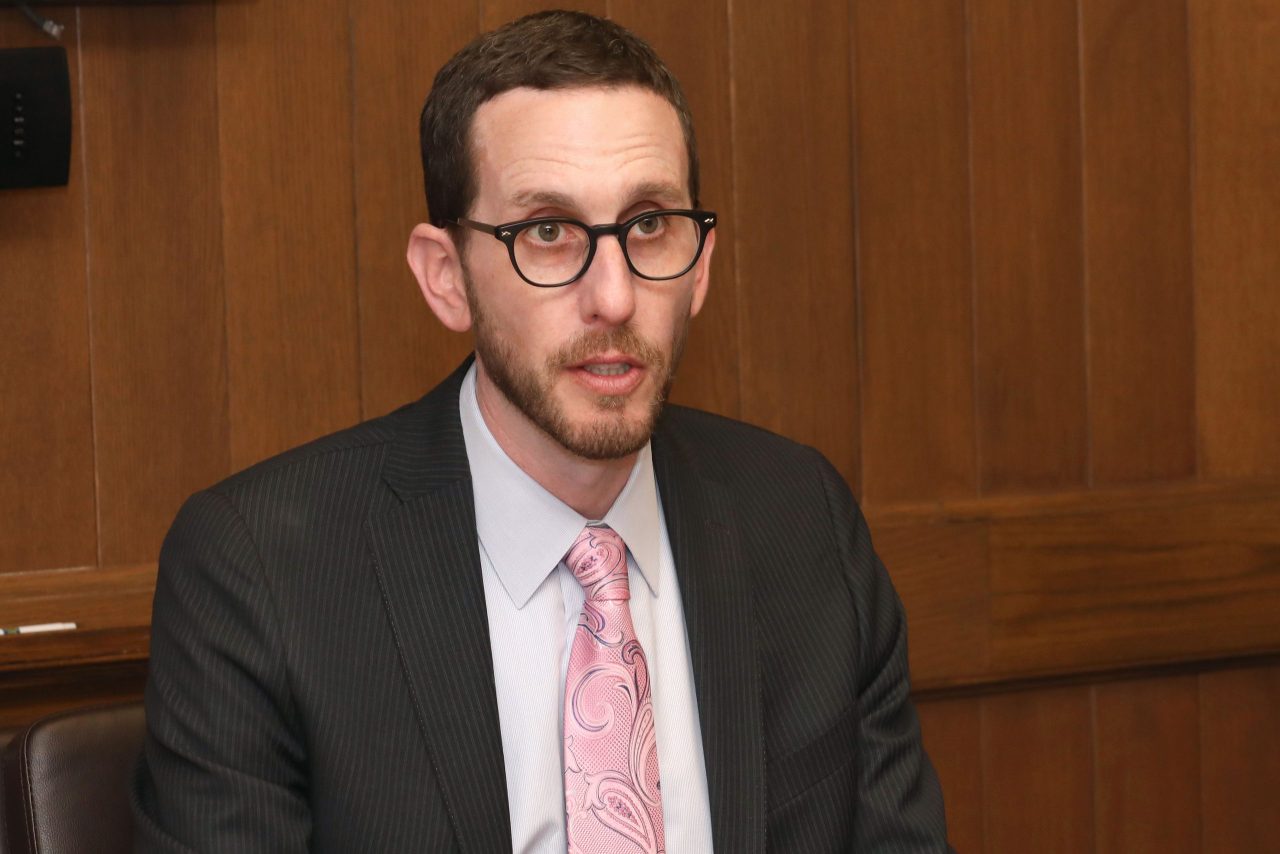 A man in a dark suit and light pink tie wearing glasses speaks.