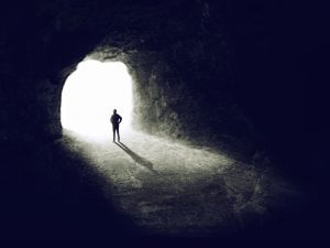 A man stands with his hands on his hips in front of a beam of light that is coming in from an entrance of a dark tunnell