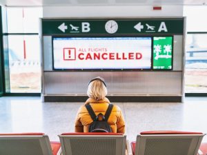 A woman wearing a yellow jacket,over-ear headphones and a backpack sits at an airport terminal and looks at a notice that reads "all flights cancelled"