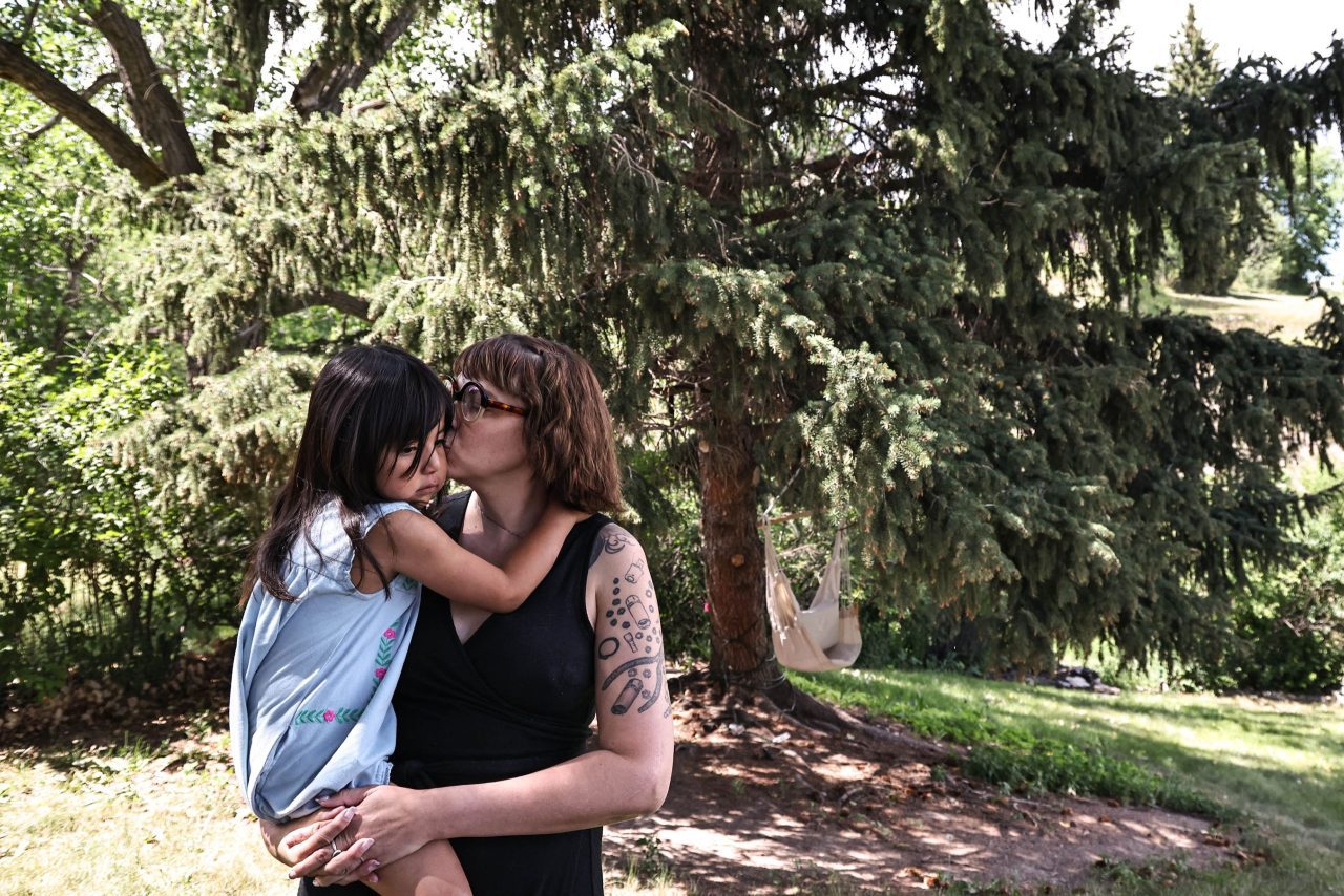 A woman holding a young child and kissing her on the cheek stands outside in front of a coniferous tree.