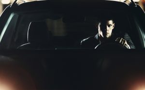 Closeup of macho looking man in a car at parking lot during night. He's staring directly at camera and looking a bit intimidating. Wearing dark suit. Lit from bot sides with sharp light. Front view.