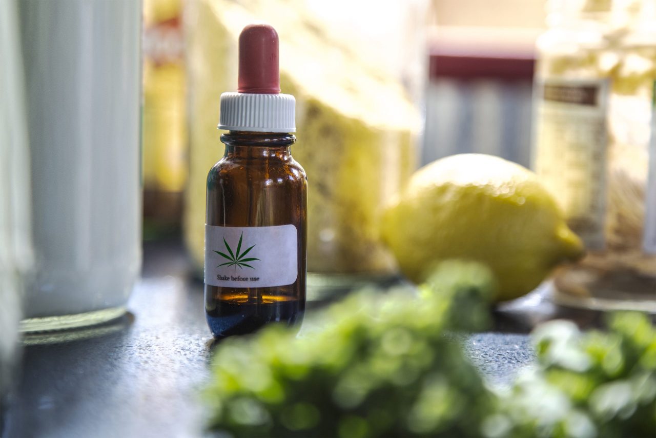 A tincture bottle of CBD oil is seen on a table. A warning on the label reads, "Shake before use."