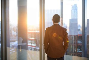 Male office worker looking at view through window of modern office over city.