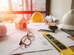 A pair of glasses, a small model home, a level tool, a measuring tape, a set of pens and two construction hats sit atop a blueprint of a house on a desk.