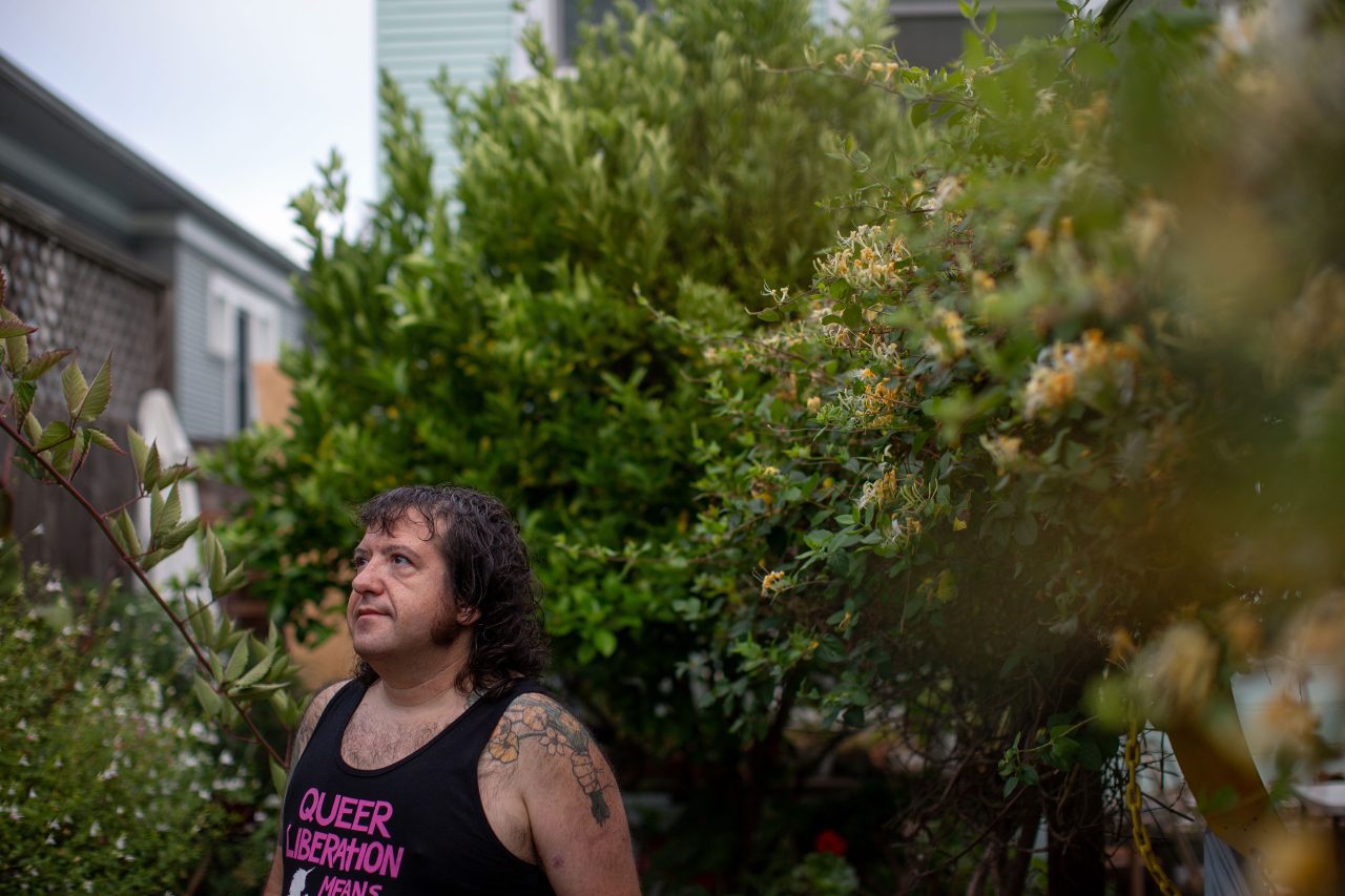 A man in a black tank-top stands outside amongst trees and bushes, looking off-camera to his right.