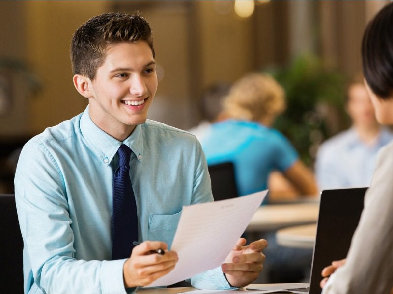 Young man in a blue button up and tie smiles while holding his resume at a table across from someone on a laptop