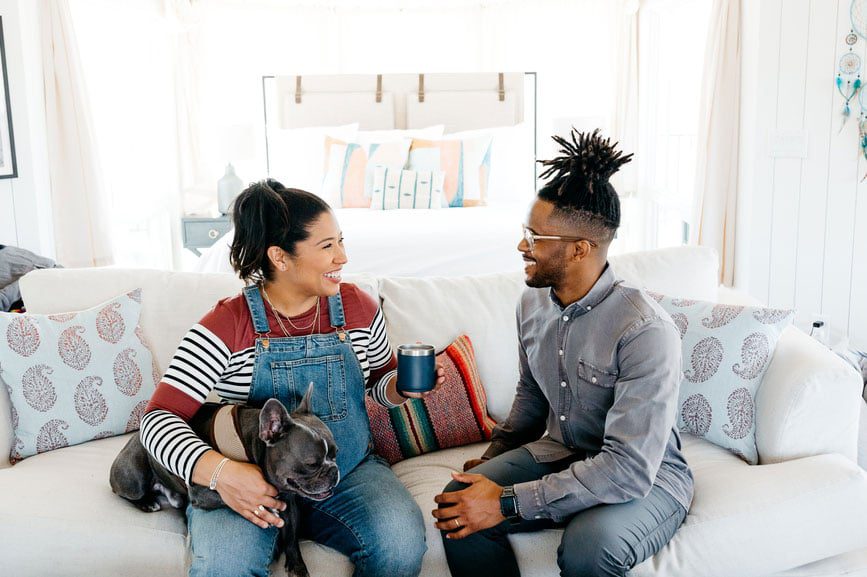 Couple sitting on a couch with their dog and smiling at eachother.