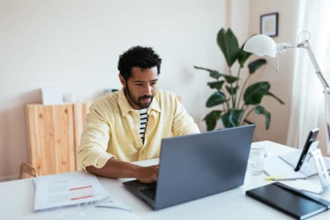 Bearded black male typing on laptop keyboard and thinking while sitting at table and working on project in light workplace