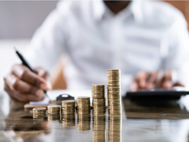 a businessman uses a calculator and a pen while sitting behind a stack of coins increasing in size