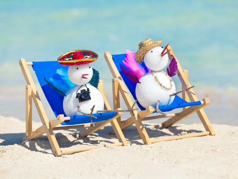 Two snowmen in the shape of birds sitting on blue beach chairs on the sand. One is wearing a sombrero and is holding a camera. The other is wearing a straw hat.