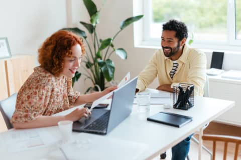 Cheerful diverse man and woman with laptop and papers smiling and working on project together while sitting at table in light workspace