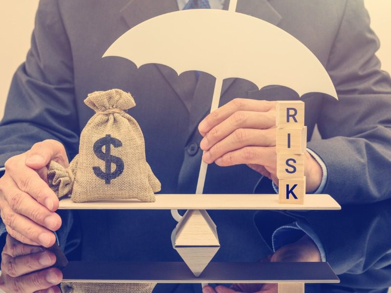 A businessman holds a white, 2 dimensional umbrella over a scale. A bag of money and wooden blocks spelling "risk" sit on either side of the scale