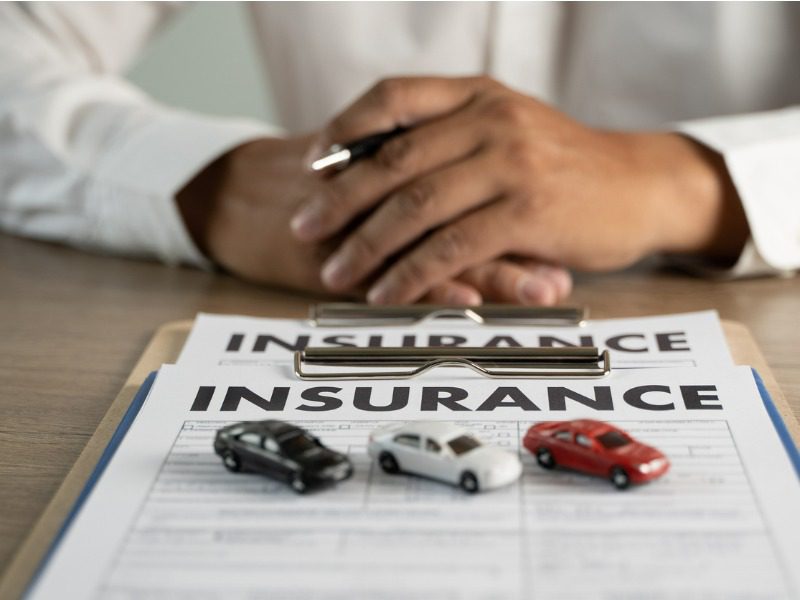 A man holding a pen sits in front of a clipboard with an insurance policy and three toy cars lined up atop it.