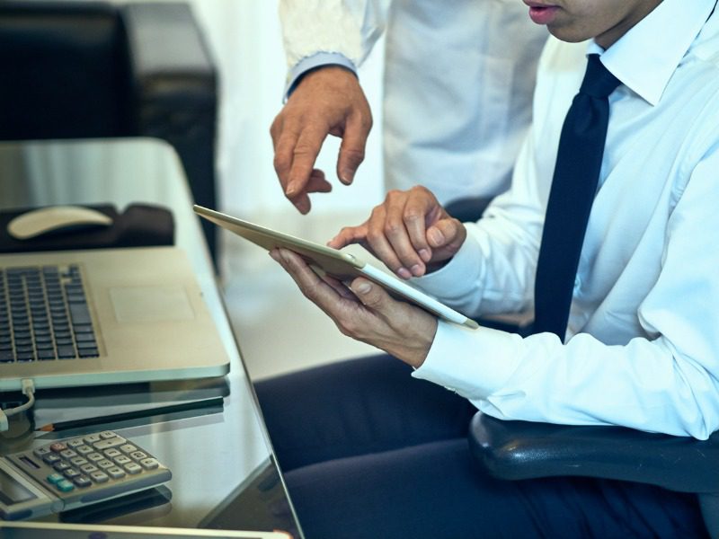 A man sits at a computer desk with a notepad in hand. Another man, both in business clothes, is leaning over his shoulder and pointing at the notepad.