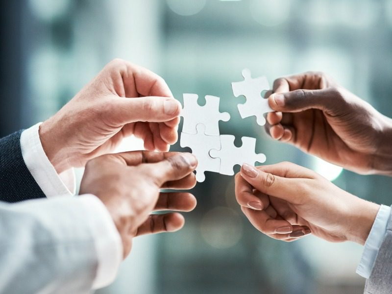 Close up shot of four businesspeople's hands. Each person is reaching forward and holding the piece of a puzzle next to their colleague's hands.