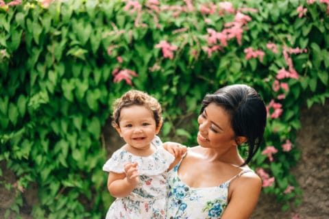 woman standing outside holding baby girl with smiles on their faces in beautiful floral sun dresses.