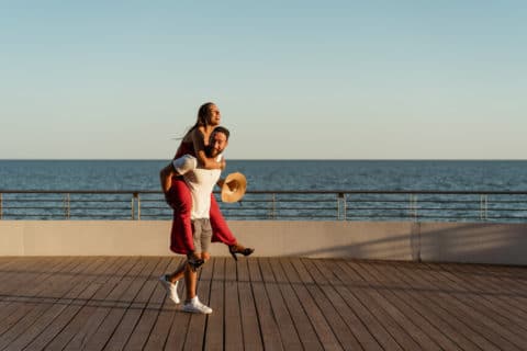 Happy man and woman doing piggyback outdoor during a walk on a waterfront in summer. Holiday time. Copy space.