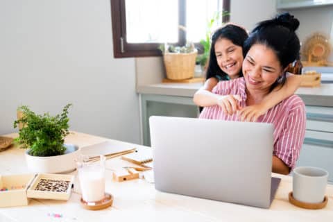 Young mother working from home using a laptop at kitchen accompanied by her daughter who hugs her and smiles
