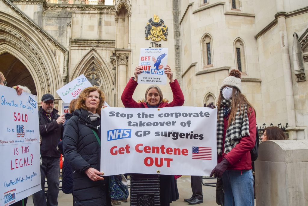 A photo shows two demonstrators holding a banner that reads, "Stop the corporate takeover of NHS GP surgeries. Get Centene out."