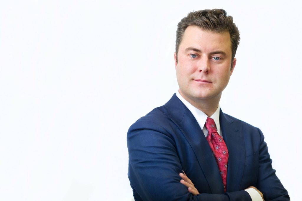 Scott Irwin, Navacord, wearing a blue suit and red tie stands with his arms crossed against a white background