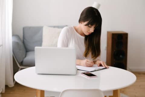 Young female entrepreneur sitting at a table with a laptop and mobile phone on it. She is writing on a clipboard