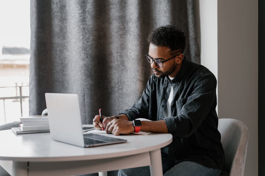 Black man in glasses sitting at table with laptop and writing in notepad while working on freelance project at home