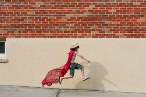 young boy wearing scarf as cape jumps into air, pretending to be superhero