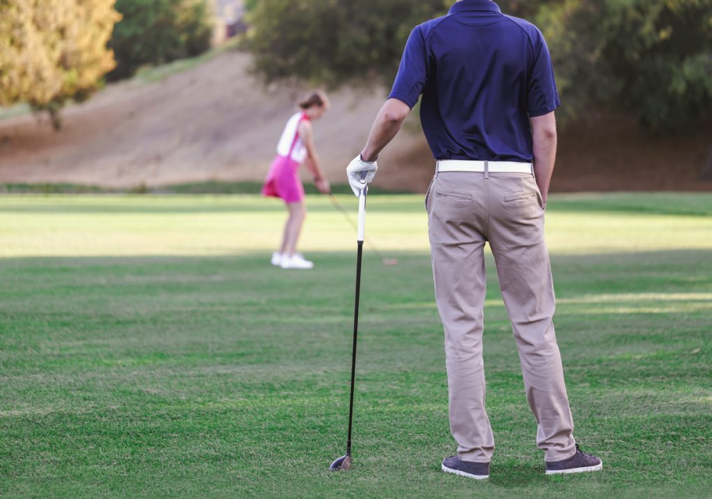 Back of man wearing golf trousers watching his wife hit a golf ball 