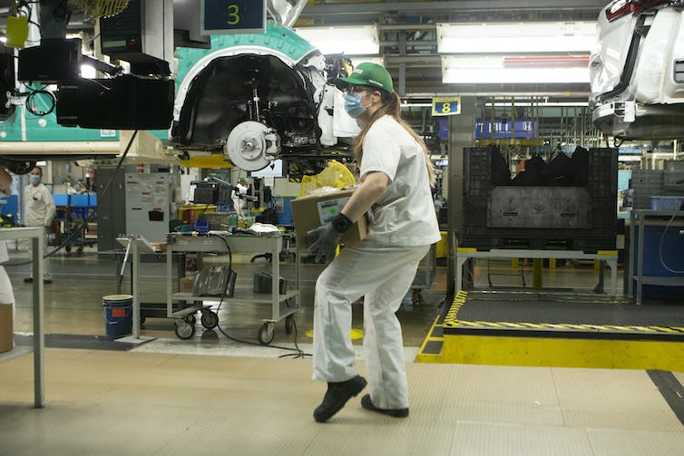 An autoworker with a ponytail, ball cap and face mask carries a box at an auto manufacturing plant.