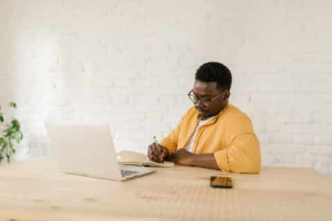 Young Afro-American man sitting at a table in his living room, writing something in his notebook from his laptop.