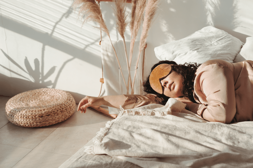 Young adult woman in pink silk pajamas sleeps on bed with blindfold over eyes in home environment in early sunny morning.