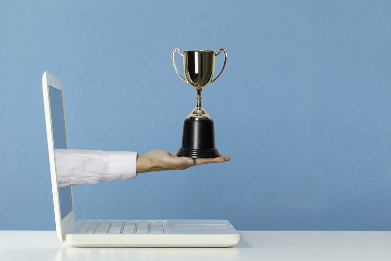 Hand holding golden trophy award is reaching out of white laptop screen on white table in front of blue wall. Representing winner and first price of competition.