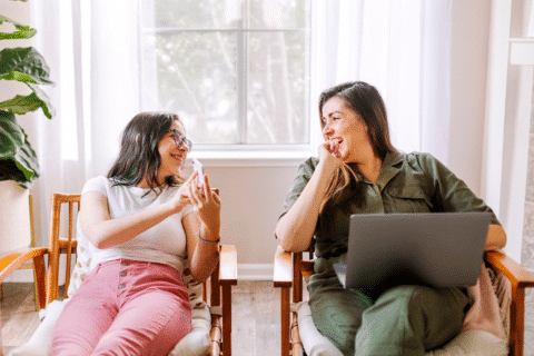 Daughter sharing her phone's screen to mother sitting on rattan lounge chairs in a bright living room