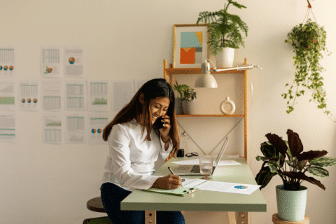 Side view of beautiful administrative woman calling by phone happily while taking notes at small cozy workspace